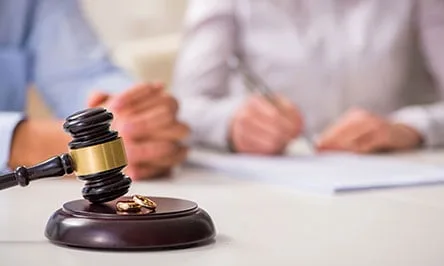 A judge’s gavel and two wedding rings on a desk, with two people in the background reviewing or signing documents.
