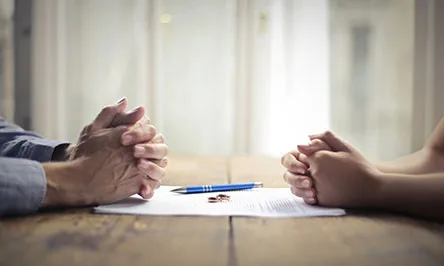 Two people sit across from each other at a table with a document, pen, and two rings placed between them.