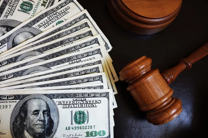 A wooden judge’s gavel rests beside several one hundred dollar bills spread out on a dark surface.