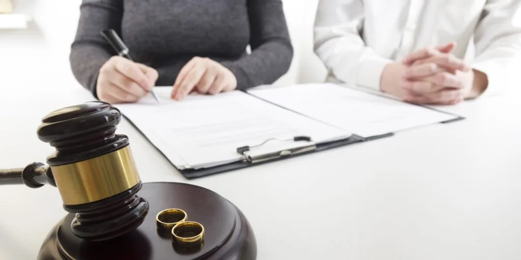 A gavel and two rings sit on a table in front of two people reviewing paperwork, suggesting a legal proceeding related to marriage or divorce.